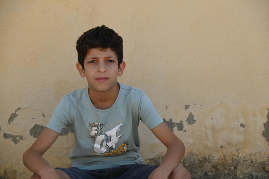 An adolescent boy looks toward us. The wall behind him has peeling paint. 