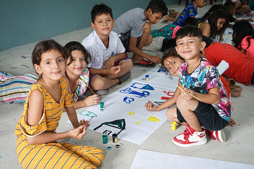 Children sit on the floor and smile as they participate in an art activity.