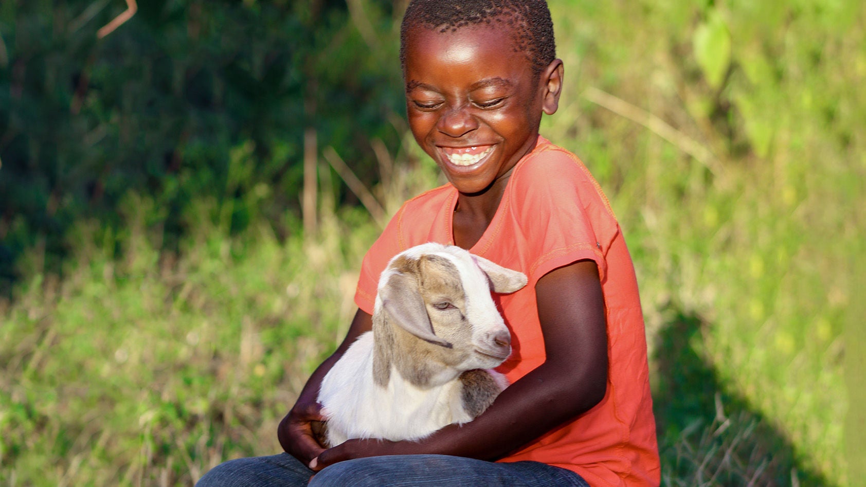 A boy in a red shirt sits outdoors, holding a goat. (Zambia, 2021)