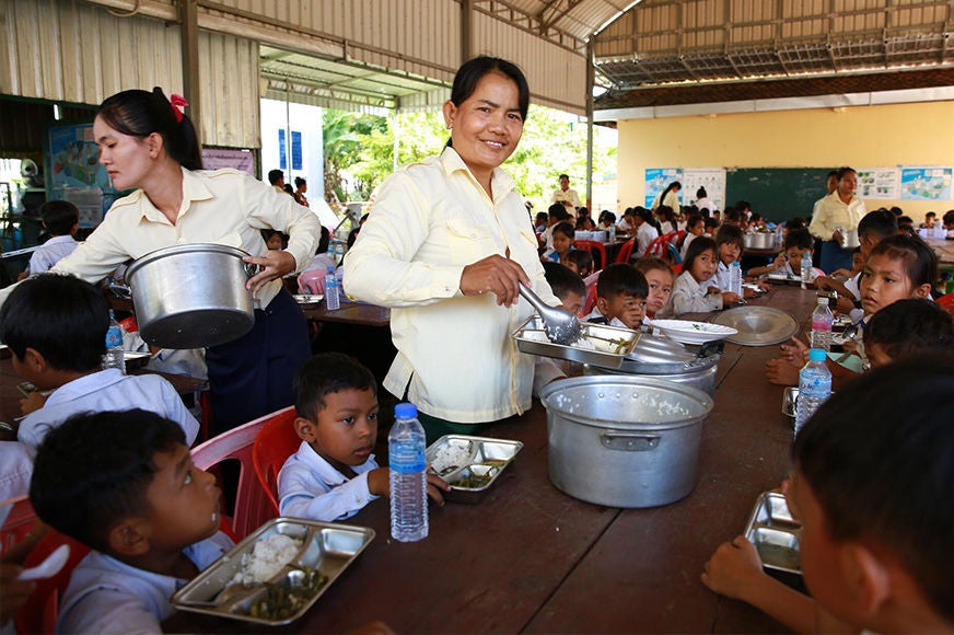 A woman holding a ladle distributes meals to students in a school feeding program, which aims to improve nutrition, food access, and education.