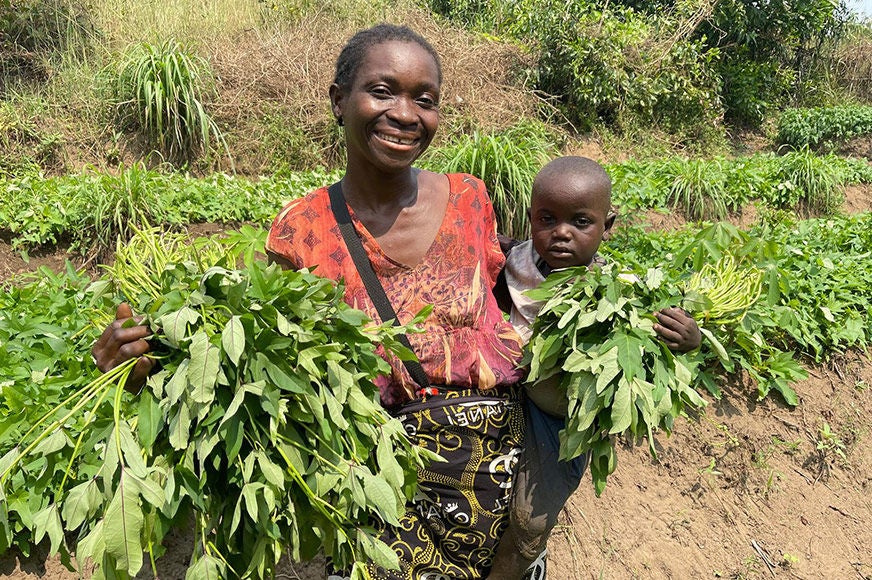 A woman is outside pulling vegetable crops while holding her baby on her hip. (Democratic Republic of Congo, 2024)