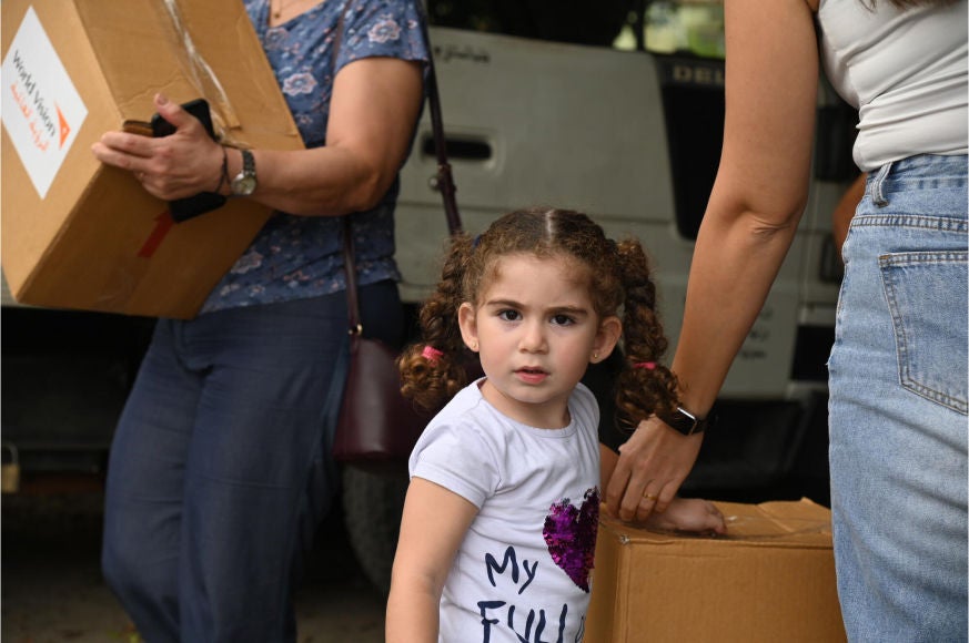 A school-age girl looks straight to camera as she stands amidst boxes being unloaded from a truck. 