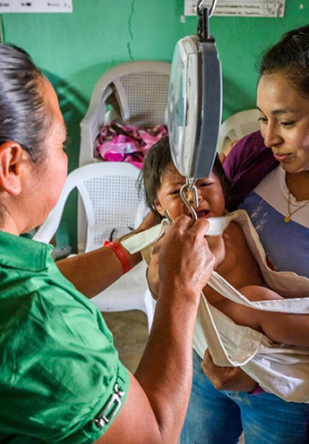  Baby, carried by their mother, being weighed on a scale by health worker.