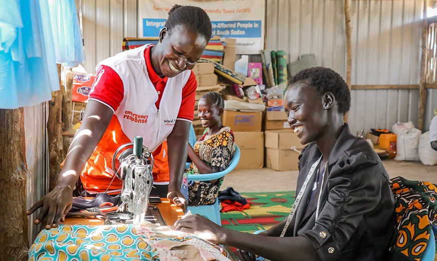 A World Vision volunteer helps Yar (right) as she sews clothes at a centre for displaced women and girls. (South Sudan, 2022)