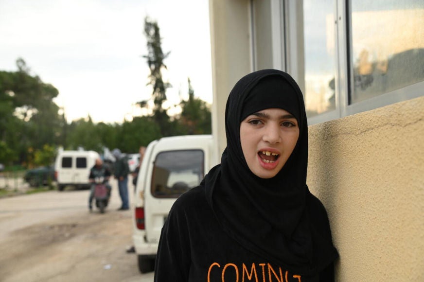 A school-aged girl wearing a head scarf leans against a wall, looking toward us. 