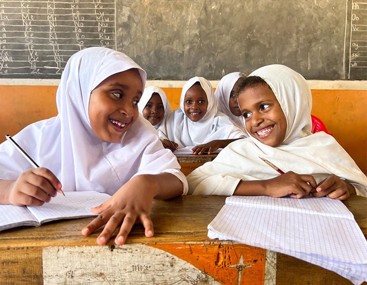 Two students are smiling at each other while sitting at a desk inside a classroom.