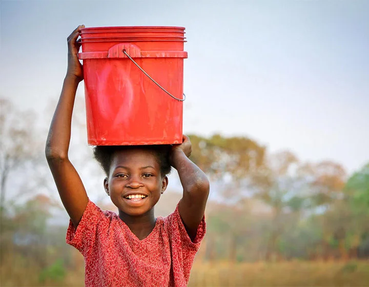 A child smiling as she holds a bucket over her head.