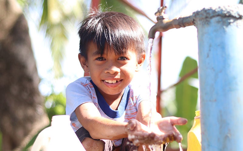 A little boy smiles as he washes his hands at an outdoor faucet.