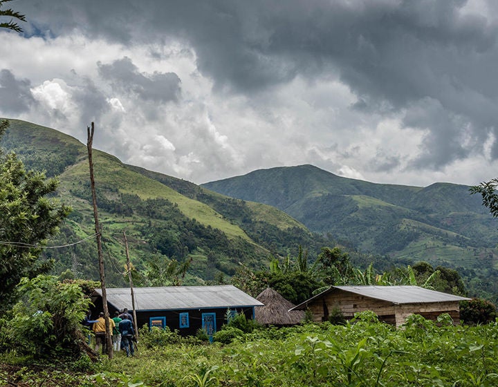 A wide shot image of a green forested area in the Democratic Republic of Congo. In the distance there are people walking towards two large cabins. (Democratic Republic of Congo, 2022)