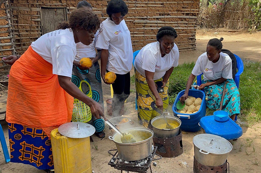 Five women stir food in pots and prepare an outdoor meal together. (Democratic Republic of Congo, 2023) 