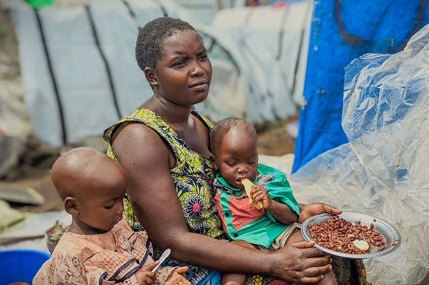 A mother sits outside their shelter with her two children while feeding them. (Democratic Republic of Congo, 2024)