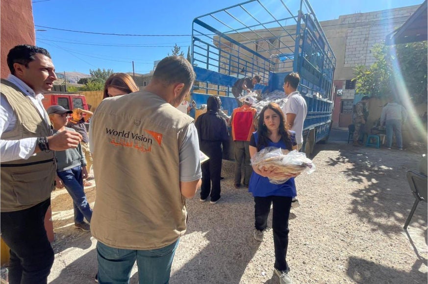 A man in a World Vision vest looks at his clipboard. Around him, people carry food supplies to their shelters. 