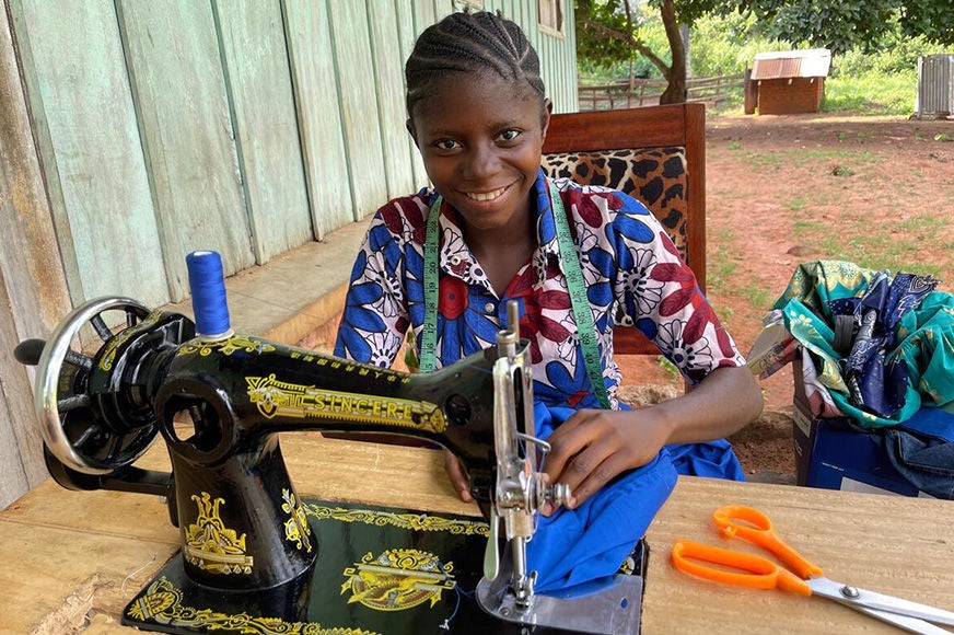 A smiling teenage girl is sewing clothes using a machine that World Vision provided her. (Democratic Republic of Congo, 2023)