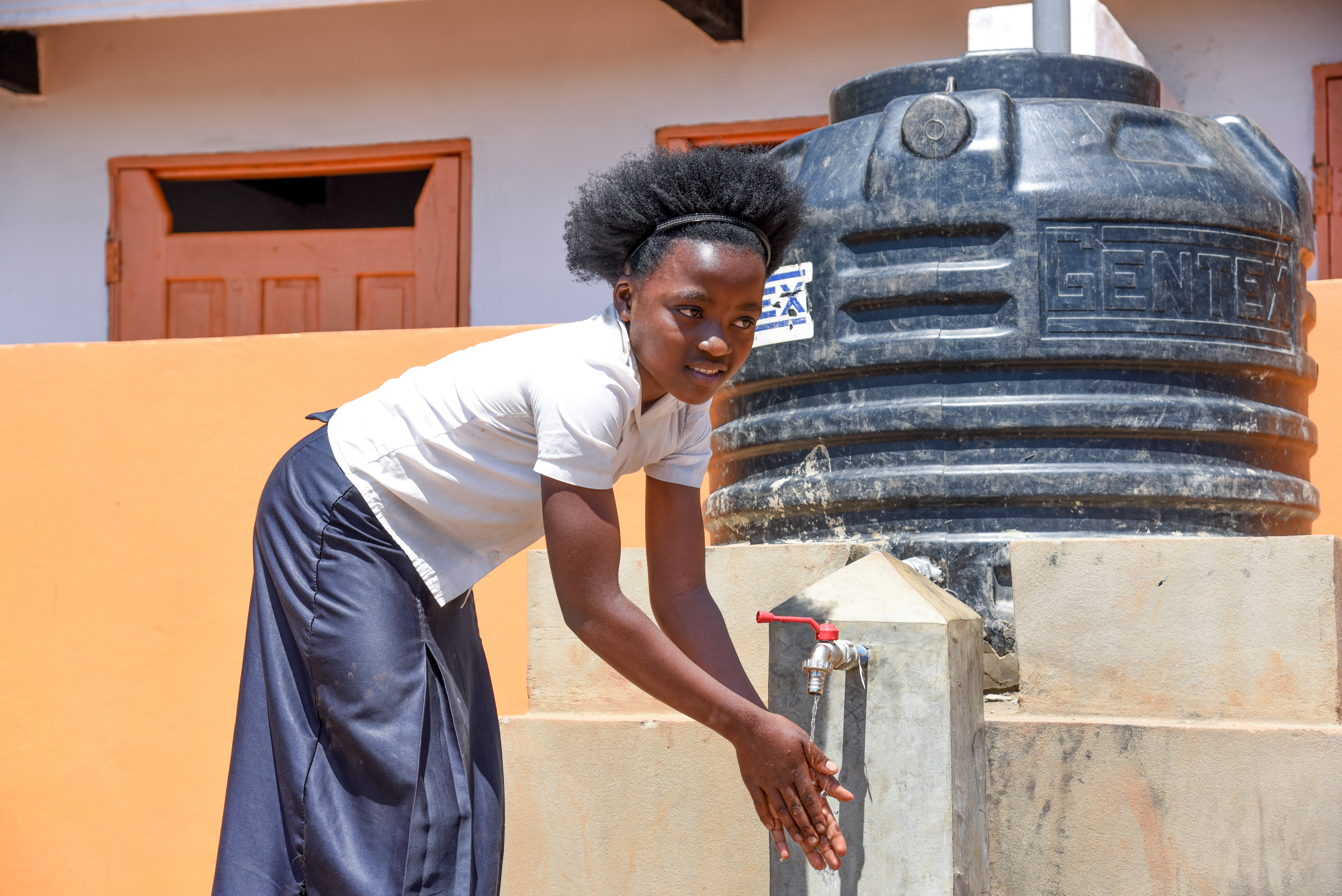 An adolescent girl leans over a tap and spout, washing her hands in clean water.