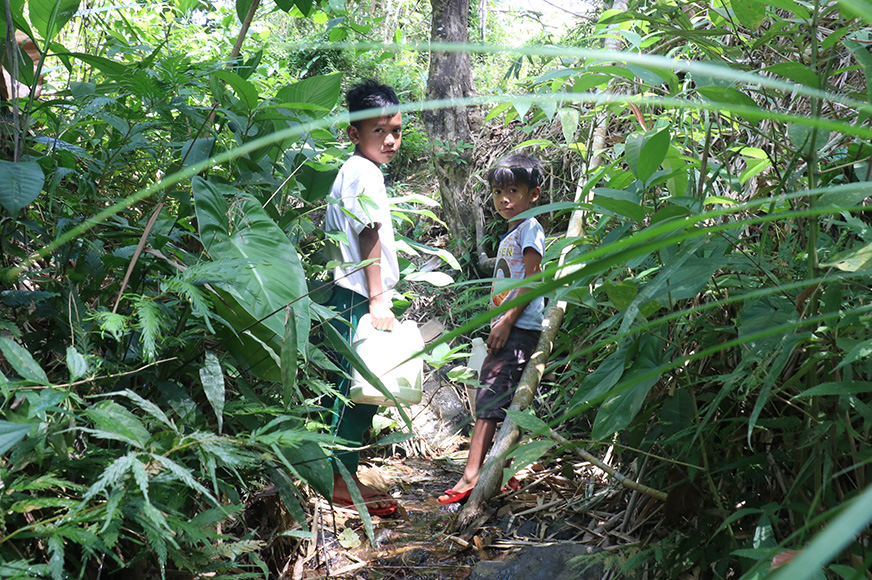 Jory (right) and his classmate, holding empty water containers, look back through overgrown brush in a forest. (Philippines, 2020)